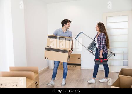 Young couple of man holding box and Woman with ladder and tool bag standing in apartment with carton boxes smiling and looking at each other Stock Photo