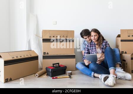Young cute couple sitting with tablet and smiling on floor with carton boxes and tools in cozy apartment Stock Photo