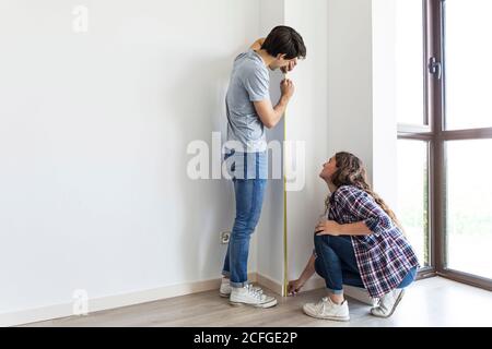 Young male and female measuring wall with linear tape in apartment with carton boxes Stock Photo