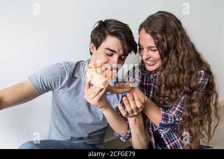 Cute young couple sitting on floor eating pizza at carton box near ladder and laughing Stock Photo