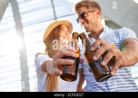 Attractive young man and Woman smiling and looking at each other while clinking bottles of cold beer in roofed pathway on sunny day Stock Photo
