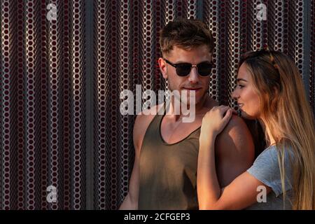 Handsome young man and pretty Woman smiling and looking at each other while standing near metal wall on street and embracing Stock Photo