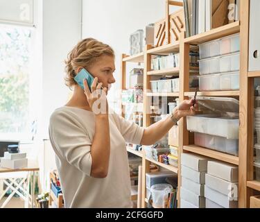 Manufacture of glass products, workpiece. Young female entrepreneur speaking on mobile phone and looking on shelves, happy lady in sunny day in workshop. Women's small business Stock Photo