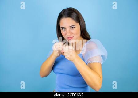 Young beautiful woman over isolated blue background Punching fist to fight, aggressive and angry attack, threat and violence Stock Photo