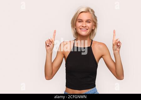 Freestyle. Young blonde woman in top standing isolated on white pointing up at copy space laughing happy Stock Photo