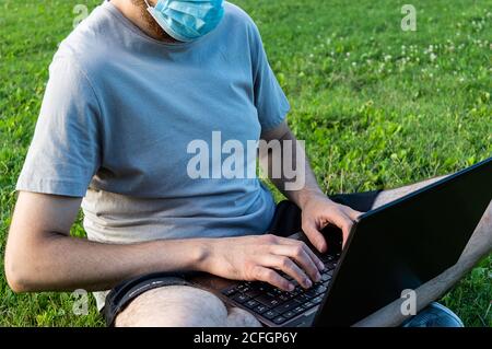 A caucasian bearded man is sitting on the grass in the sun with his laptop. Wearing surgical mask, prevent coronavirus, smart working on nature Stock Photo
