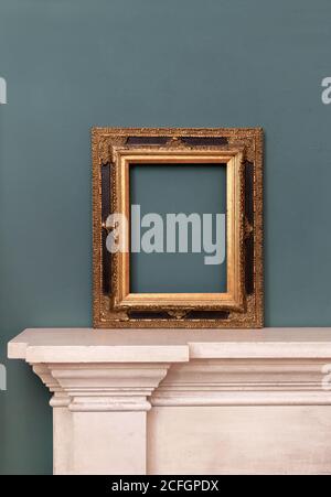 Gilded or golden empty rectangular vintage frame on a mantelpiece for a picture or painting leaning against a green wall Stock Photo