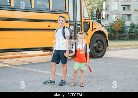 Happy Caucasian brothers students near yellow school bus. Smiling kids holding hands and going back to school in September. Education system and learn Stock Photo