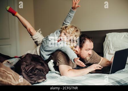 Work from home with kids children. Father working on laptop in bedroom with child daughter on his back. Funny candid family moment. New normal during