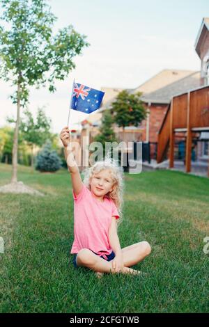 Happy Caucasian blonde girl holding Australian flag. Smiling child sitting on grass in park waving Australia flag. Kid celebrating Australia Day Stock Photo