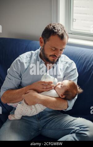 Middle age Caucasian father feeding newborn baby from bottle with milk. Man parent holding rocking child on his hands. Authentic lifestyle candid real Stock Photo