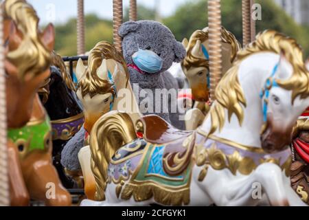 A teddy bear toy wearing a protective mask seats on a carousel horse in a city during the restrictions with the novel coronavirus COIVD-19 pandemic Stock Photo