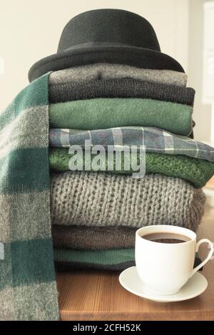 Pile of green and grey sweaters with a black hat on top and a hot drink white cup on the side, background with copy space, cozy, hygge or seasonal war Stock Photo