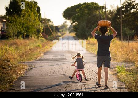 Little child girl, riding a bike, with her young father carrying a big halloween pumpkin over his head, on a country road at sunset. Back view. Stock Photo