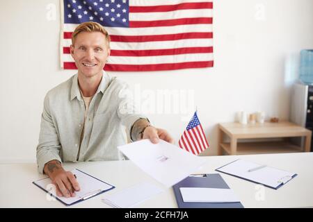 Portrait of smiling young man handing papers to people while registering voters at polling station on election day, copy space Stock Photo