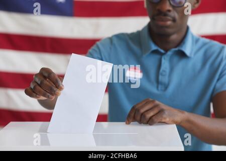 Cropped portrait of African-American man putting vote bulletin in ballot box while standing against American flag on election day, copy space Stock Photo