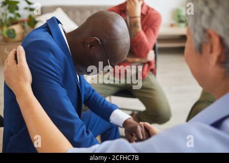 Side view portrait of grieving African-American man crying in support group with mature woman comforting him, copy space