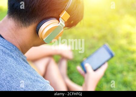 A Man wear headphone and play phone. He sitting on the green grass. Stock Photo