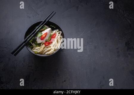 Spicy asian soup with chicken bouillon and meat, red chilli pepper, fresh cilantro and onion, japanese noodles in black soup bowl with chopsticks on b Stock Photo