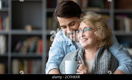 Smiling young man hugging mature mother drinking tea Stock Photo