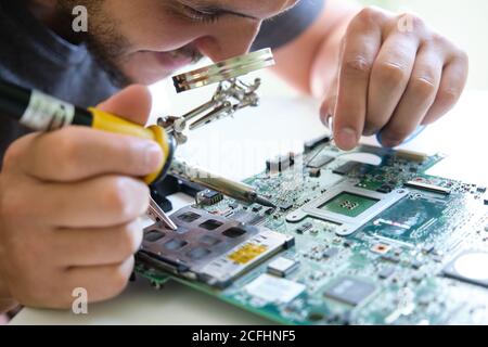 Young adult repairing a printed circuit board with a soldering iron through a magnifying glass. Technology concept. Stock Photo