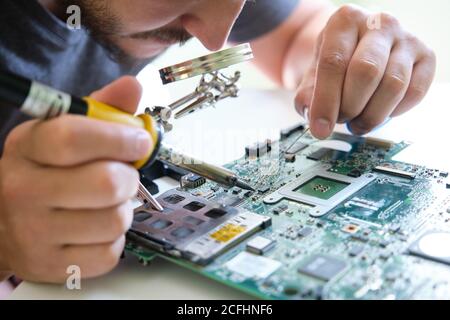 Young adult repairing a printed circuit board with a soldering iron through a magnifying glass. Technology concept. Stock Photo