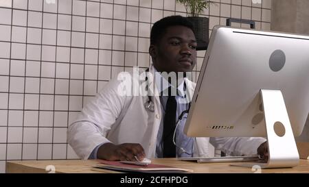 Entering african male doctor sitting down at desk and start working. Stock Photo