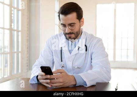 Focused doctor wearing glasses using smartphone, sitting at work desk Stock Photo