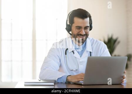 Smiling young doctor in headset consulting patient online, using laptop Stock Photo
