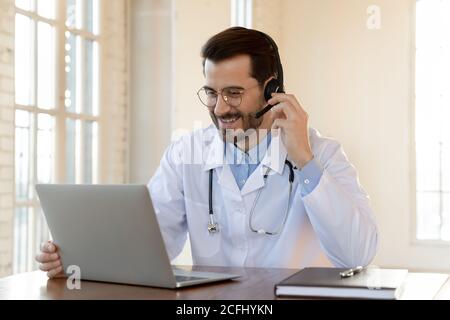 Smiling doctor wearing headset working on laptop, consulting patient Stock Photo