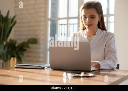 Focused young woman doctor working on laptop online in office Stock Photo