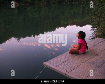 Little girl sitting on a pontoon by the river and contemplating the sky of a sunset Stock Photo