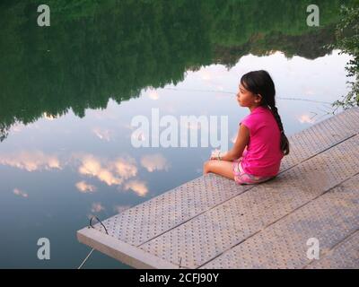 Pensive little girl seen in profile sitting on a pontoon by the river, contemplating nature Stock Photo