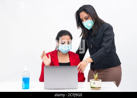 Two professional Indian women working on computer while wearing Covid protection mask, work from home, Corporate employees discussing project Stock Photo