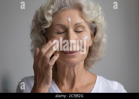 Close up satisfied middle aged woman applying moisturizing face cream Stock Photo