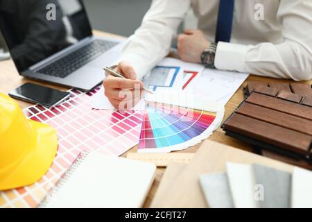 Man sits at table holding pen in his hands on table lie smartphone laptop color palette and a yellow hard hat. Stock Photo