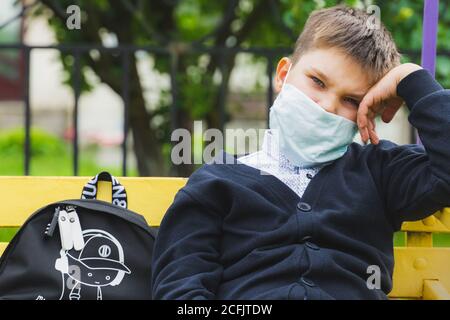 tired kid boy in mask looking to camera. sad schoolboy looking sitting on a bench  with a backpack in the school yard waiting for the bus. Back to sch Stock Photo