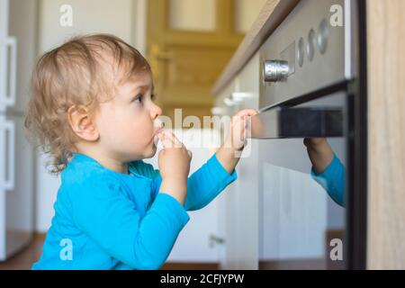 Small and curious child playing with the knobs of the oven in the kitchen. Danger for unattended children, accident prevention at home conceptual phot Stock Photo