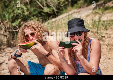 Company of friends sitting on tree log in forest and eating delicious watermelon while enjoying summer holiday together Stock Photo