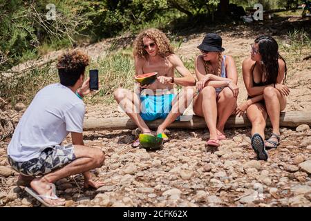 Group of friends in summer wear relaxing on shore in woods and cutting watermelon while enjoying holiday together Stock Photo