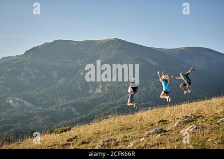 Company of delighted friends jumping on hill in mountains while enjoying freedom during summer vacation Stock Photo