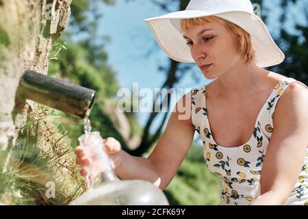 Side view of female in dress and sunhat filling plastic bottle with fresh water from natural spring in rock in sunny day Stock Photo