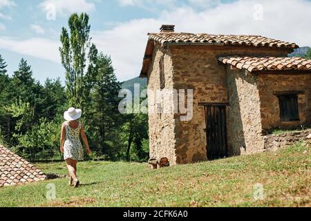 Back view of female in dress and straw hat walking near old stone house in village on sunny day Stock Photo
