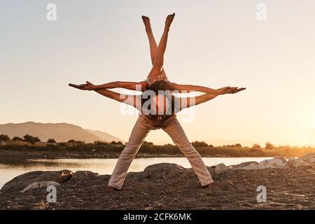 Strong man with slender woman on back balancing with raised crossed legs while practicing acro yoga during sunset in summer