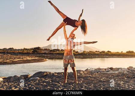 Slim woman leaning on shoulder of man and balancing on arm while doing acroyoga together near river