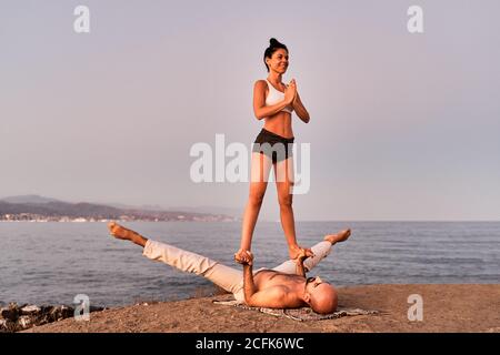 Slim woman in Mountain pose with Namaste gesture balancing on hands of strong man while doing acroyoga at seaside