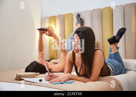 Busy female students lying on bed and preparing for exam while using smartphone and taking notes in workbook Stock Photo