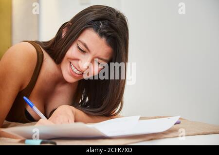 Cheerful female student lying on bed at home and writing on paper while preparing homework assignment Stock Photo