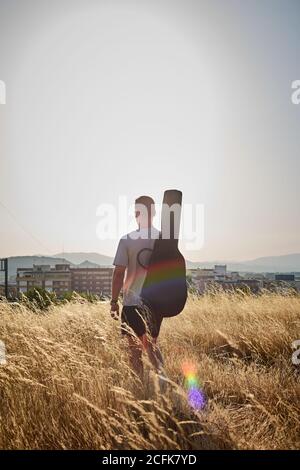 Back view of male musician with guitar in case walking along dried field in summer and looking away Stock Photo