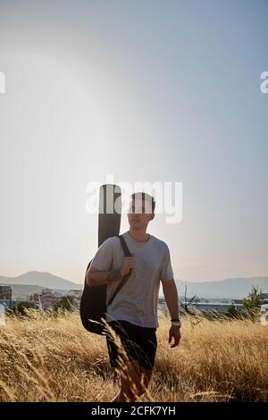 Peaceful male musician with guitar in case walking along dried field in summer and looking away Stock Photo